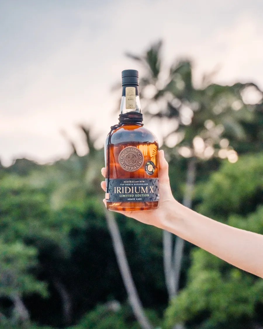 The current Iridium X Agave Cask bottle held up against Far North Queensland rainforest at dusk. The front label reads Australian Rum, Far North Queensland, Iridium X, Limited Edition, Agave Cask