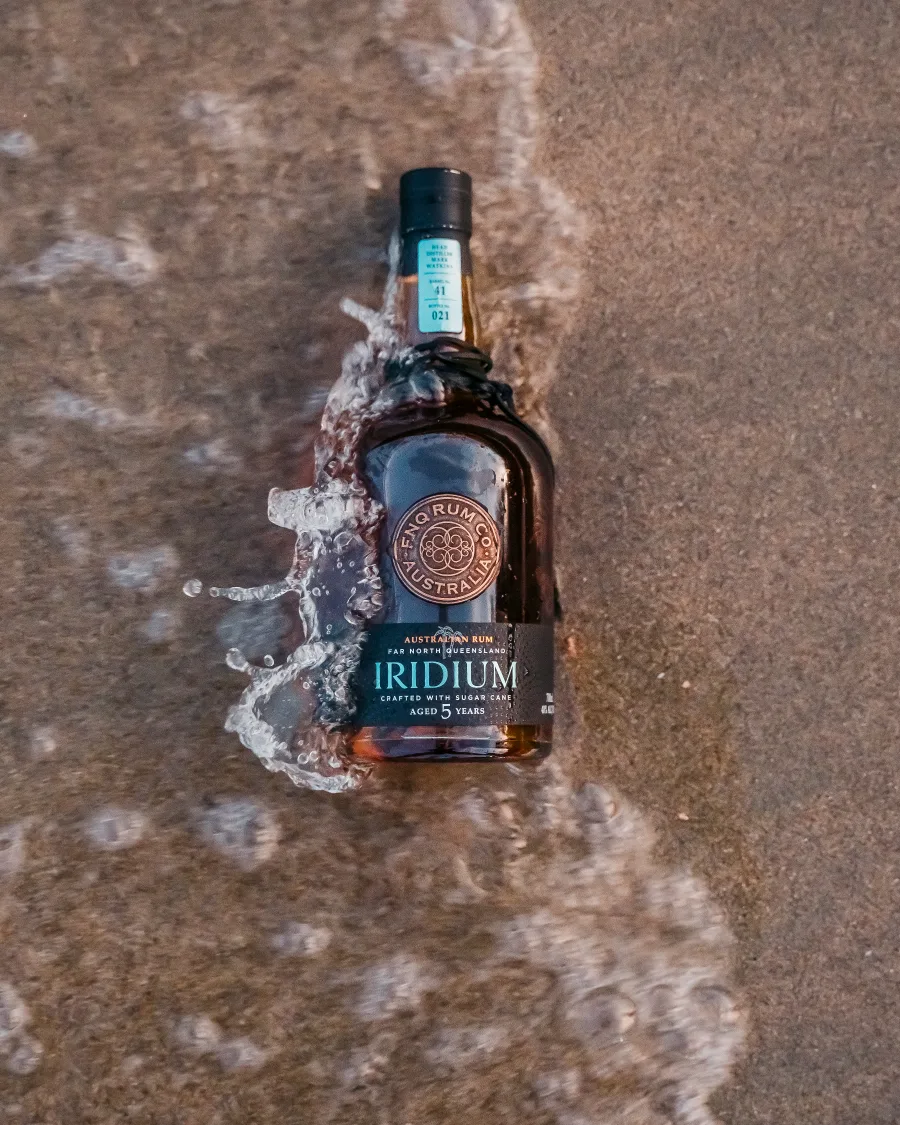 A bottle of FNQ Rum Co. Iridium lying in shallow water at the edge of a Far North Queensland rock pool, its neck label reading barrel 41, bottle 021