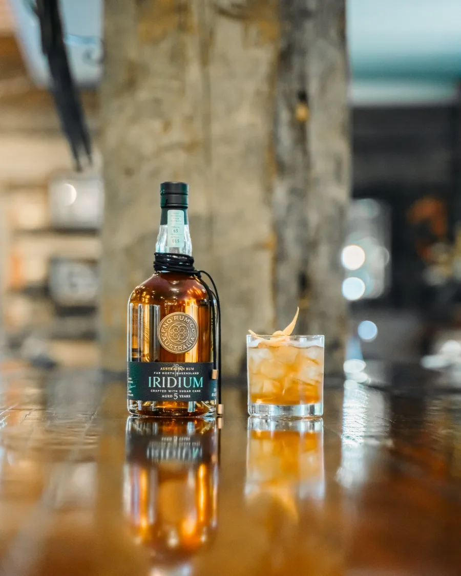 A bottle of FNQ Rum Co. Iridium beside a glass of rum over ice on the bar. The drink pictured is a plain serve, not one of the cocktails listed on this page